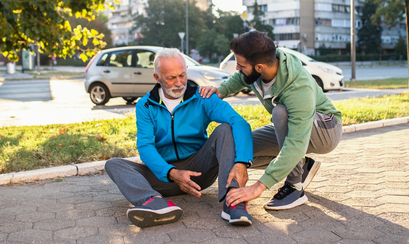 Man holding ankle sitting on sidewalk with another man assisting him